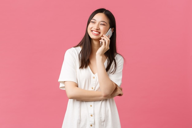 Belleza, emociones de las personas y concepto de tecnología. Amistosa niña bonita sonriente en vestido blanco hablando por teléfono feliz, mirando hacia arriba y de pie fondo rosa alegre.