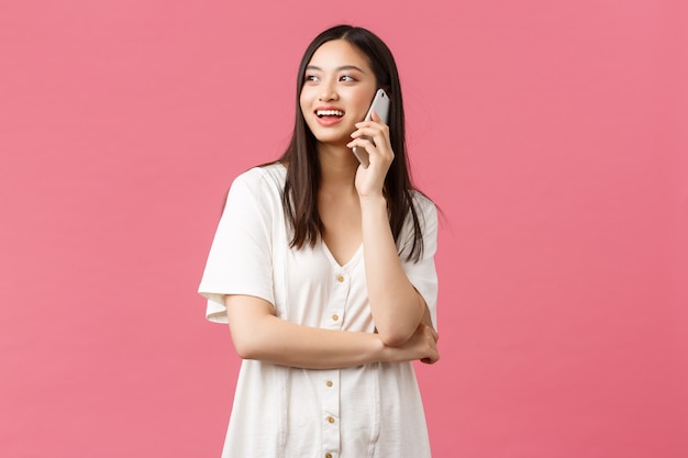 Belleza, emociones de las personas y concepto de tecnología. Amistosa niña bonita sonriente en vestido blanco hablando por teléfono feliz, mirando hacia arriba y de pie fondo rosa alegre.
