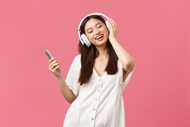 Belleza, emociones de las personas y concepto de tecnología. Chica asiática bonita y despreocupada que disfruta de la música en auriculares inalámbricos, cierra los ojos y sonríe escuchando su canción favorita, sosteniendo el teléfono inteligente y bailando.