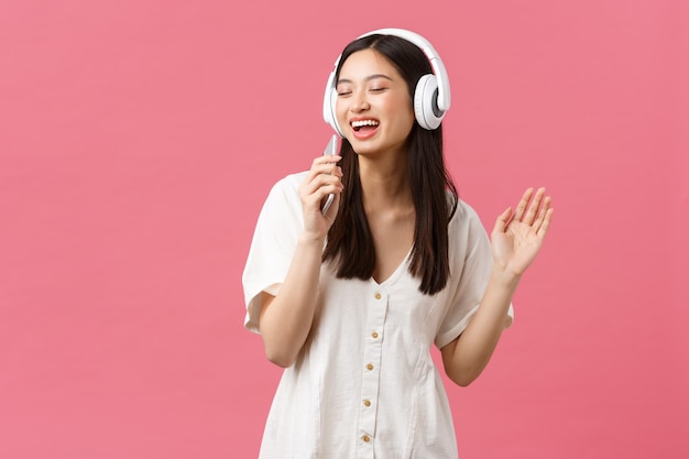 Belleza, emociones de las personas y concepto de tecnología. Chica asiática feliz despreocupada que usa la aplicación de karaoke del teléfono móvil, cantando en el micrófono del teléfono inteligente, escucha música en auriculares, fondo rosa