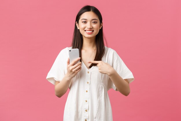 Belleza, emociones de las personas y concepto de tecnología. Chica asiática guapa complacida con vestido blanco apuntando con el dedo al teléfono inteligente como una aplicación increíble, recomiendo la aplicación de teléfono móvil