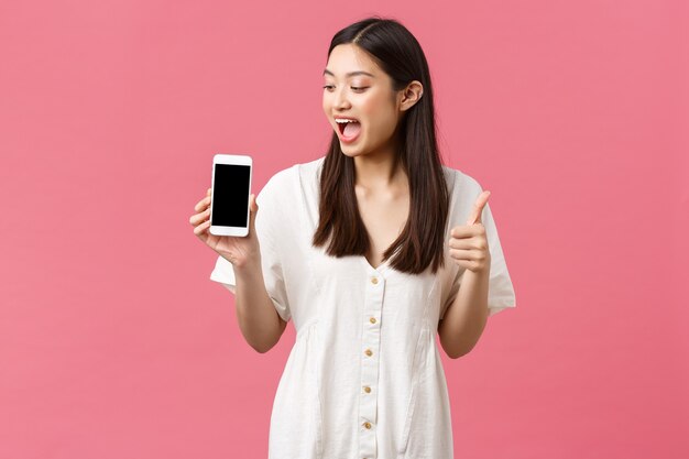 Belleza, emociones de las personas y concepto de tecnología. Emocionada chica asiática feliz, blogger demuestra una aplicación impresionante, mostrando la pantalla del teléfono móvil y el pulgar hacia arriba en regocijo, fondo rosa.