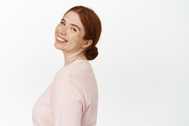 Belleza y estilo de vida de las mujeres. Retrato de una joven pelirroja sonriendo, girando la cabeza hacia la cámara con expresión sincera y feliz, sonriendo despreocupada, de pie contra un fondo blanco.