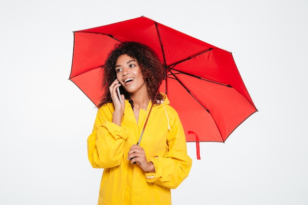 Belleza feliz mujer africana en gabardina escondiéndose bajo el paraguas mientras habla por el teléfono inteligente y mirando a otro lado sobre fondo blanco.