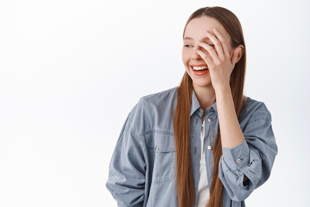 Belleza Hermosa y elegante adolescente tocando la piel facial limpia e hidratada mirando a un lado el espacio de copia de texto promocional y sonriendo de pie contra el fondo blanco