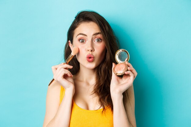 Belleza. Labios fruncidos de chica glamour tonta, aplicando maquillaje con pincel y mostrando rubor en la cámara, de pie contra el fondo azul.