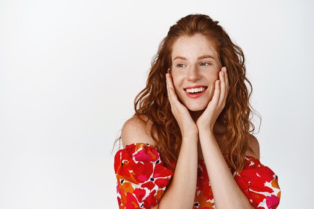 Belleza Linda mujer pelirroja sonriente en vestido floral de verano mirando coqueta a un lado y sonriendo de pie feliz contra el fondo blanco