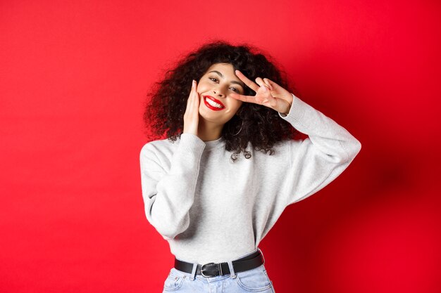 Belleza y maquillaje. Mujer joven feliz con el pelo rizado, tocando la cara y mostrando el signo v con una linda sonrisa, de pie contra el fondo rojo.