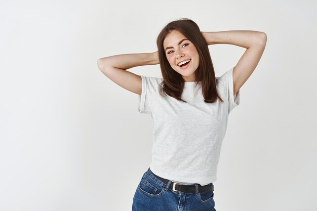 Belleza. Mujer joven feliz y relajada mirando despreocupada al frente, tomados de la mano detrás de la cabeza, apoyada contra la pared blanca