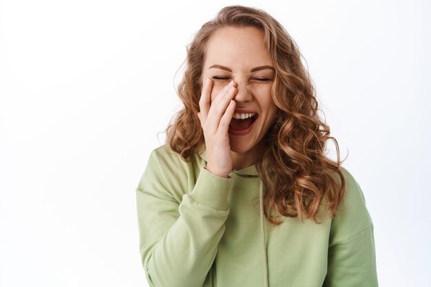 Belleza Mujer joven feliz tocando la cara y riendo cubriendo la mejilla y sonriendo despreocupada de pie en sudadera con capucha verde contra el fondo blanco.