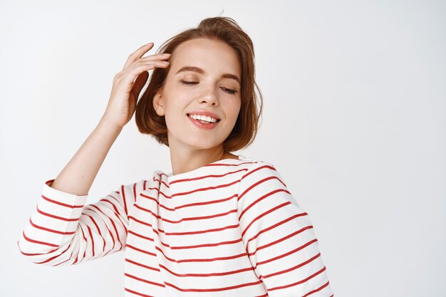 Belleza. Mujer tierna feliz con sonrisa sincera y piel fresca y natural, tocando el corte de pelo y sonriendo con los ojos cerrados, de pie contra la pared blanca