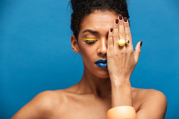 Belleza relajada mujer de raza mixta con cosméticos coloridos en la cara posando en la cámara cubriendo un ojo con la mano, aislado sobre la pared azul