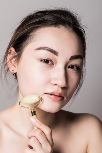 Belleza rosa jade piedra cara rodillo para terapia de masaje facial aislado en la pared gris. Retrato de mujer asiática.