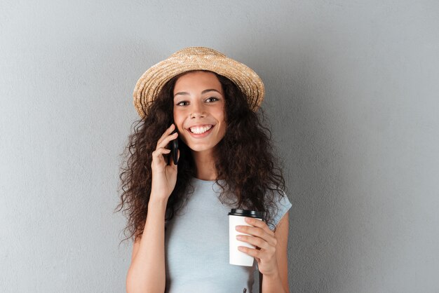 Belleza sonriente mujer rizada en sombrero hablando por el teléfono inteligente con café en mano