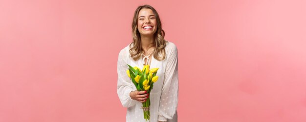 Belleza de vacaciones y concepto de primavera Despreocupada feliz atractiva chica rubia en vestido blanco sosteniendo tulipanes amarillos y riendo con expresión complacida de pie fondo rosa tener cita romántica