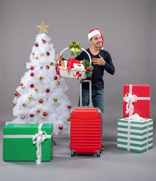 Bendito joven con gorro de Papá Noel sosteniendo una cesta de regalo con ambas manos cerca del árbol de Navidad en gris