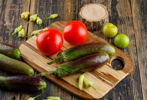 Berenjenas con tomates, pimientos, ciruelas verdes, madera en madera y tabla de cortar, vista de ángulo alto.