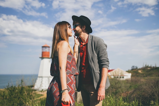 Besos estilo indie de pareja joven inconformista enamorados caminando en el campo, faro en el fondo