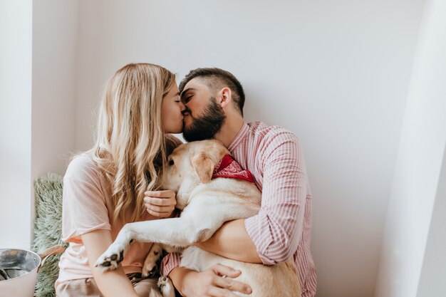 Besos de pareja romántica en la habitación soleada y luminosa. Chico con camisa rosa y su novia abrazando a un perro.