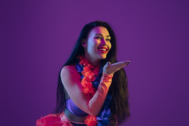 Besos sensuales. Modelo morena hawaiana en pared violeta en luz de neón. Mujeres hermosas en ropas tradicionales sonriendo y divirtiéndose. Vacaciones brillantes, colores de celebración, festival.