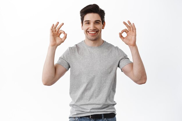 Esto esta bien. Un chico guapo sonriente muestra signos de bien, tranquiliza y da garantías de que todo lo bueno, alaba el excelente trabajo, de pie sobre el fondo blanco.