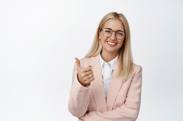 Bien hecho. Joven empresaria, empresaria en traje y gafas muestra los pulgares hacia arriba y sonríe complacida en blanco