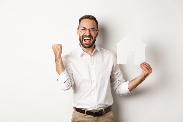 Bienes raíces. Hombre satisfecho regocijo de fundar un apartamento perfecto, sosteniendo el modelo de la casa de papel, de pie