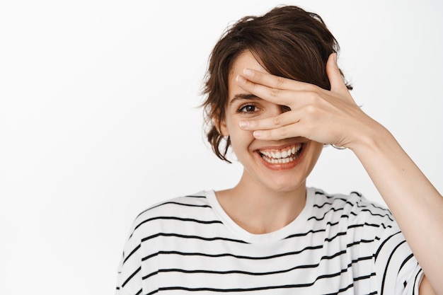 Bienestar y belleza de la mujer. Linda mujer morena sonriente que cubre los ojos, mirando con un ojo a través de los dedos, de pie sobre fondo blanco.