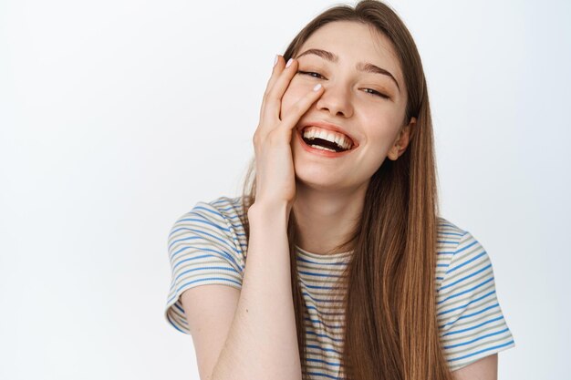 Bienestar de la mujer. Cerca de una hermosa chica natural, tocando la piel limpia y brillante, riendo y sonriendo, mostrando dientes blancos, parada sobre el fondo del estudio, aislada.