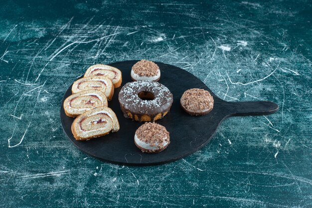 Bizcochos, rosquilla y pastel de rollo en rodajas sobre la tabla de cortar sobre la superficie azul