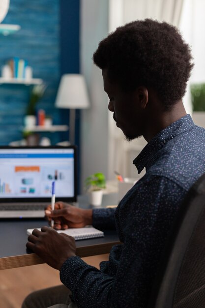 Black student writing financial strategy on notebook working at business presentation using laptop computer in living room. Teenager having online school during coronavirus lockdown