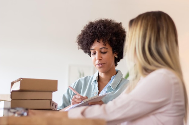 Blackwoman discutiendo el paquete con un colega y tomando nota en el portapapeles. Trabajadores de almacén preparando mercancías para su envío. Concepto de logística