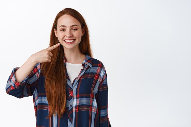 Blanqueamiento y estomatología. Chica adolescente pelirroja sonriente señalando sus dientes blancos perfectos, recomienda clínica dental, de pie en camisa a cuadros contra fondo blanco