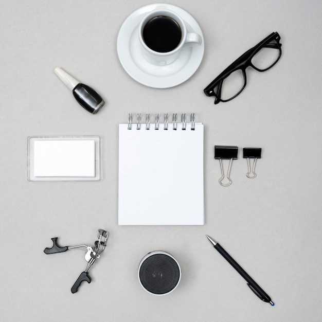 Bloc de notas blanco en blanco rodeado de una taza de café; esmalte de uñas; rizador de pestañas; altavoz; Pluma y clips de papel sobre fondo gris
