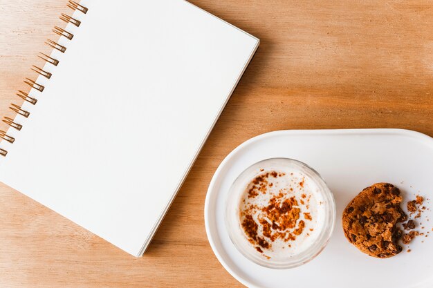 Bloc de notas en blanco espiral y vaso de café con galletas comidas en mesa de madera