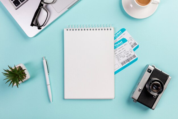 Bloc de notas de espiral en blanco con boletos aéreos rodeados con laptop, lentes, bolígrafo, cámara, taza de café en el escritorio azul