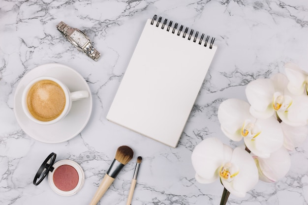 Bloc de notas de espiral en blanco; reloj de pulsera; taza de café; polvo compacto; pincel de maquillaje y flor de orquídea blanca sobre fondo de mármol con textura