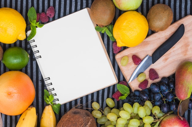 Bloc de notas de espiral en blanco rodeado de coloridas frutas y cuchillo en una tabla de cortar