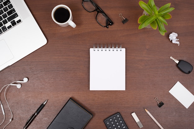 Bloc de notas espiral en blanco rodeado de suministros de oficina; gadgets Taza de té y planta en una mesa de madera