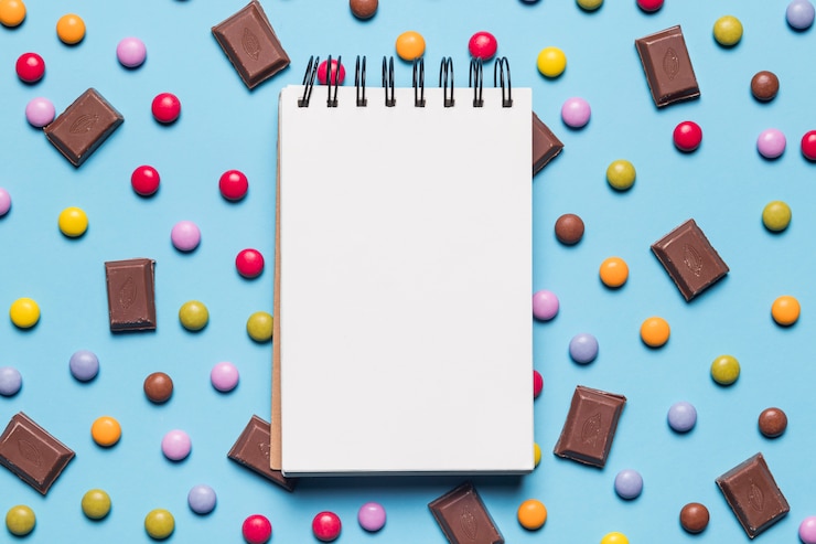 Foto gratuito bloc de notas de espiral en blanco sobre los caramelos de gema y piezas de chocolate sobre fondo azul
