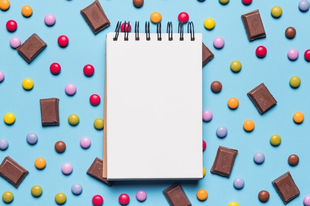 Bloc de notas de espiral en blanco sobre los caramelos de gema y piezas de chocolate sobre fondo azul