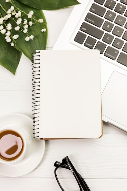 Bloc de notas espiral en blanco en la taza de café y portátil con hojas y flores en el escritorio