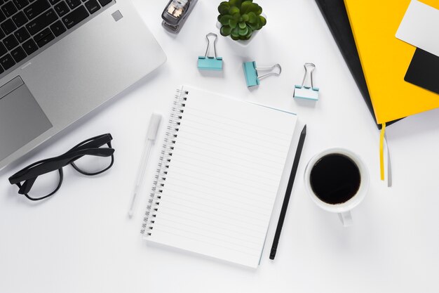 Bloc de notas espiral en blanco con taza de café y suministros de oficina en escritorio blanco
