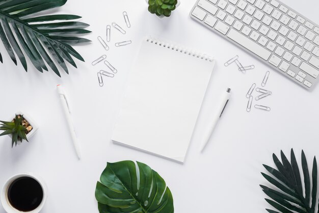 Bloc de notas espiral en blanco con teclado; Clips de papel y hojas verdes sobre fondo blanco