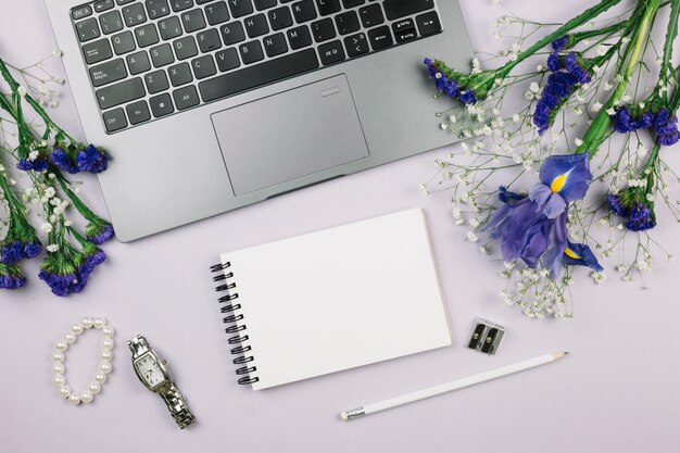 Bloc de notas en espiral; lápiz; reloj de pulsera; Pulsera y flores moradas con laptop sobre fondo blanco.