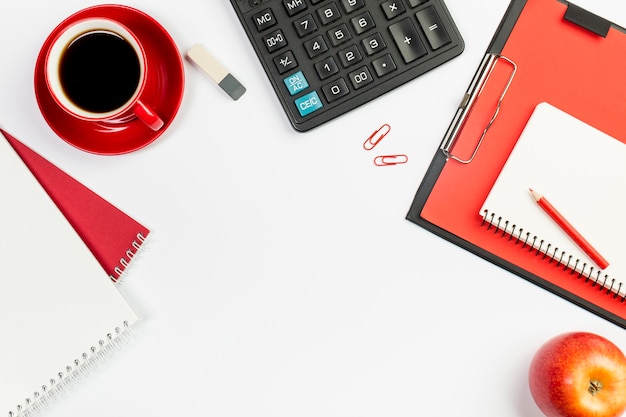 Bloc de notas en espiral, taza de café, borrador, calculadora, bloc de notas en espiral en el portapapeles con una manzana entera roja sobre fondo blanco