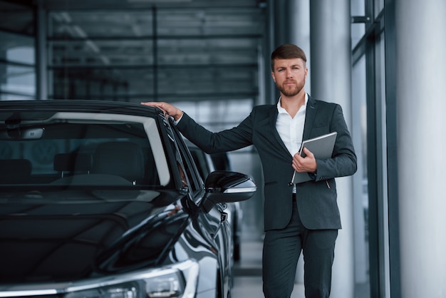 Con bloc de notas plateado en la mano. Empresario barbudo con estilo moderno en el salón del automóvil