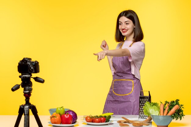 Blogger de comida linda jovencita en delantal rosa con grabación de video con verduras en la mesa