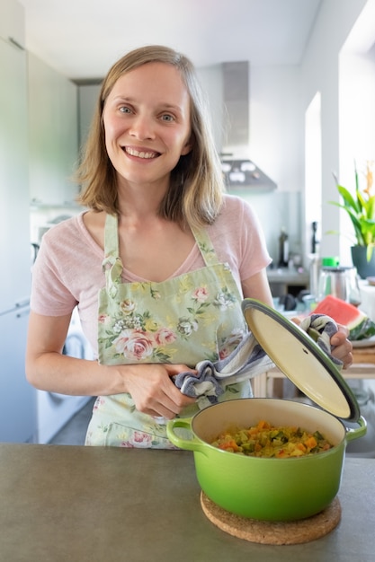 Foto gratuita blogger de comida positiva cocinando en la cocina, abriendo y mostrando una cacerola caliente con comida vegetal, mirando a la cámara y sonriendo. disparo vertical. concepto de cocina en casa