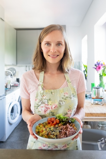 Blogger de comida con tazón de verduras. Mujer feliz con plato de verduras caseras, de pie en la cocina, mirando a cámara y sonriendo. Disparo vertical. Concepto de dieta vegetariana